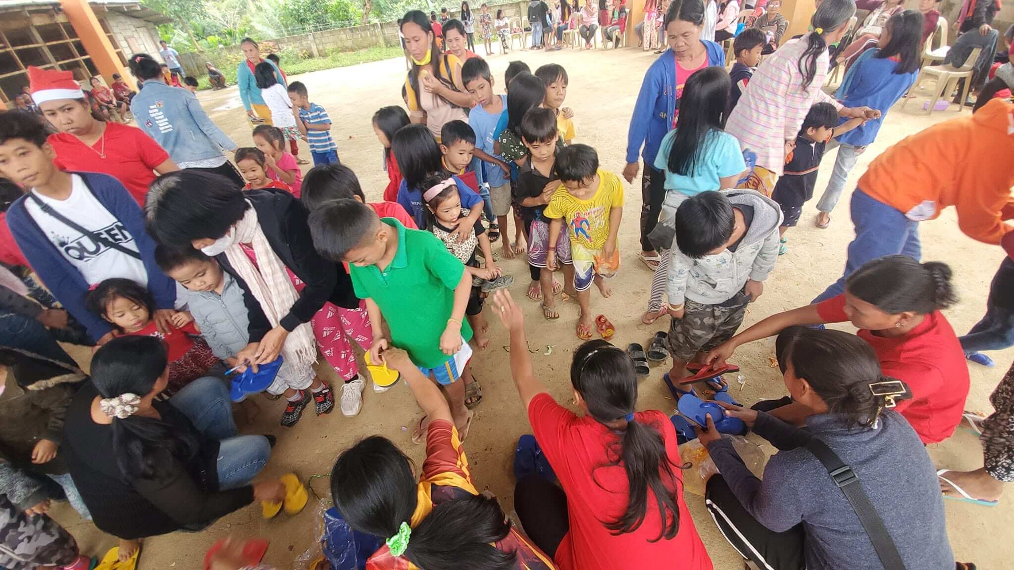 Close-up overhead view of volunteers kneeling to hand and fit slippers on children's feet as dozens of children gather tightly around the distribution area at Barangay Maasin
