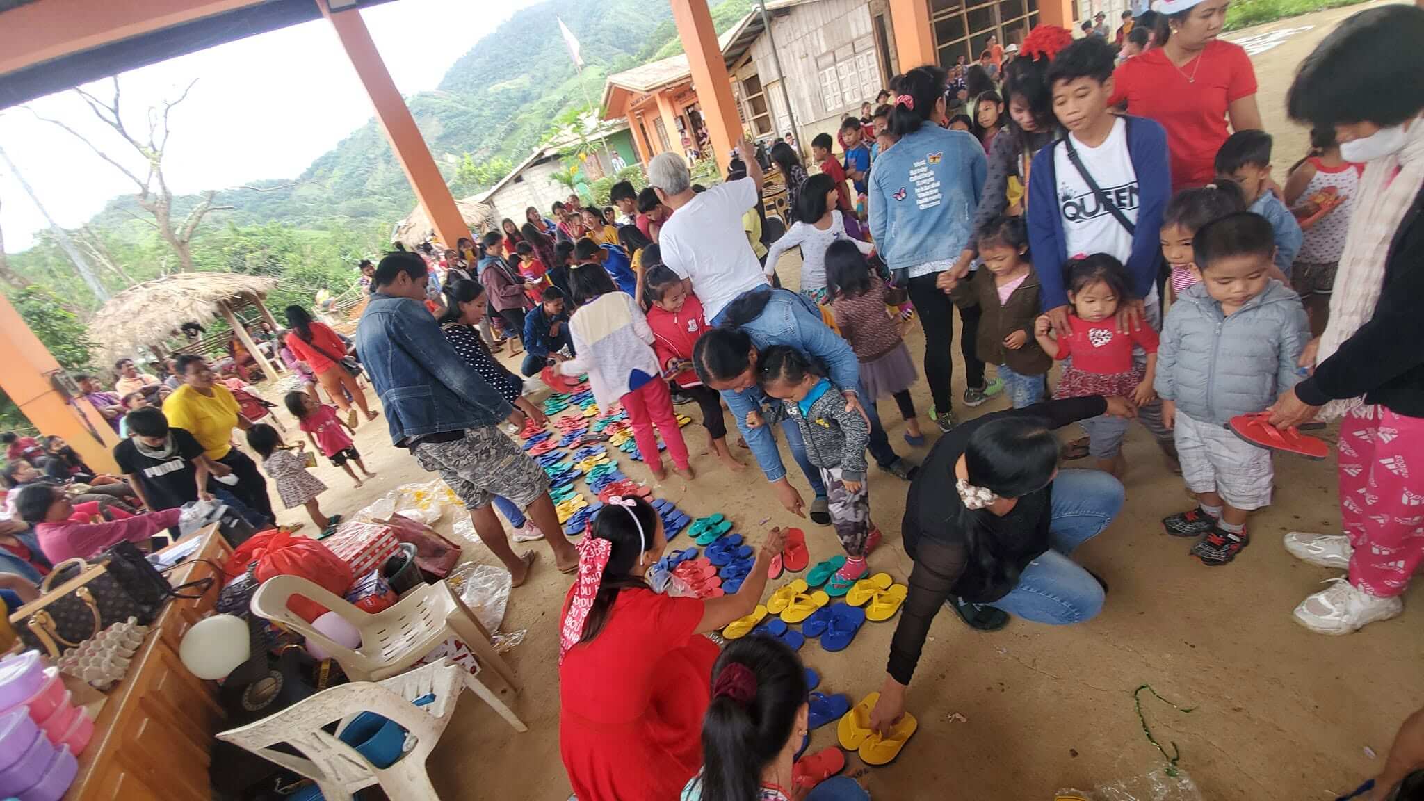 Volunteers managing children's queues and fitting slippers at the Barangay Maasin distribution area, with a large crowd of attendees visible in the background