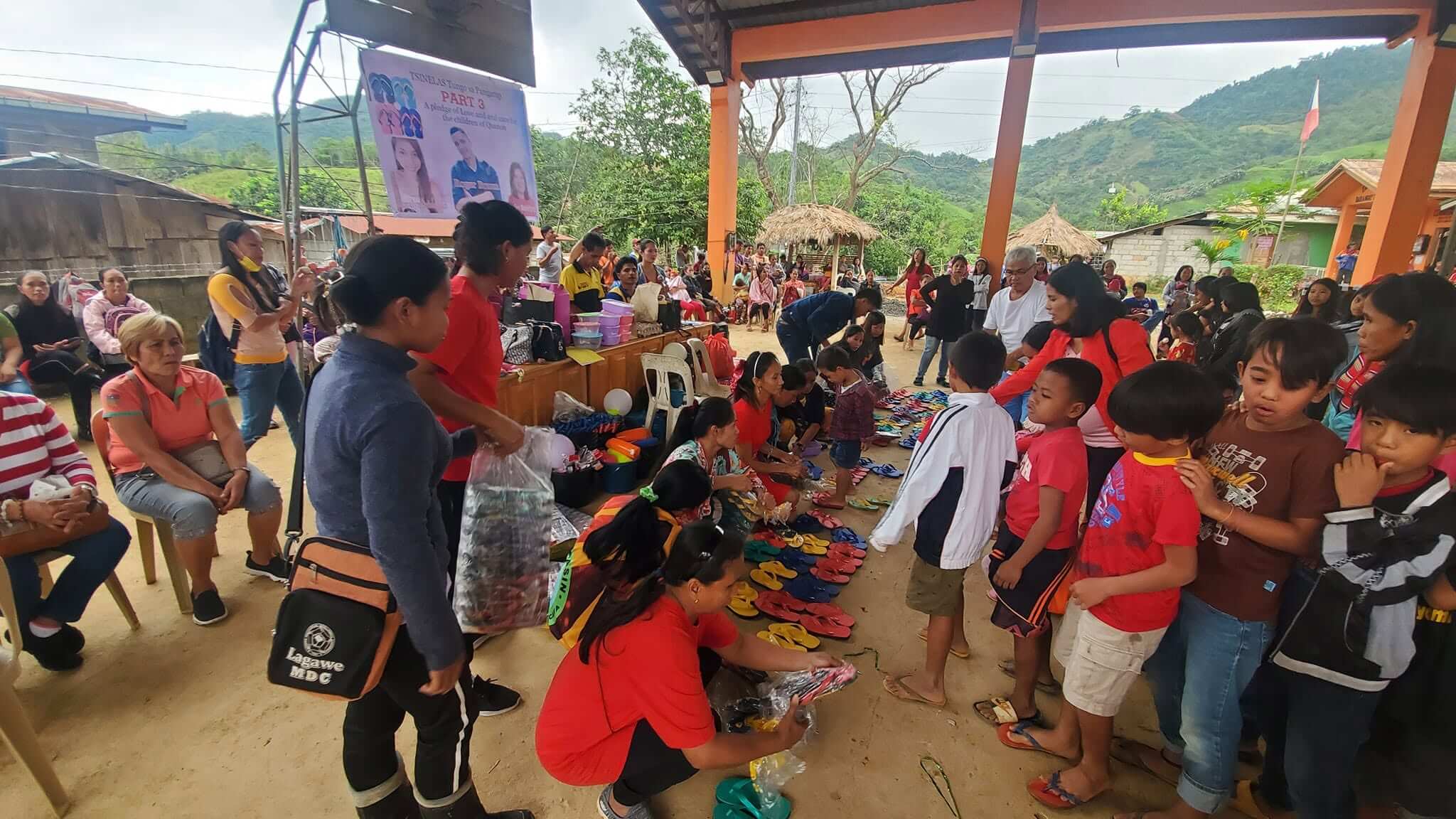 Volunteers sorting colorful slippers on the ground as children queue for distribution, the Tsinelas Tungo sa Pangarap Part 3 banner visible in the background at Barangay Maasin