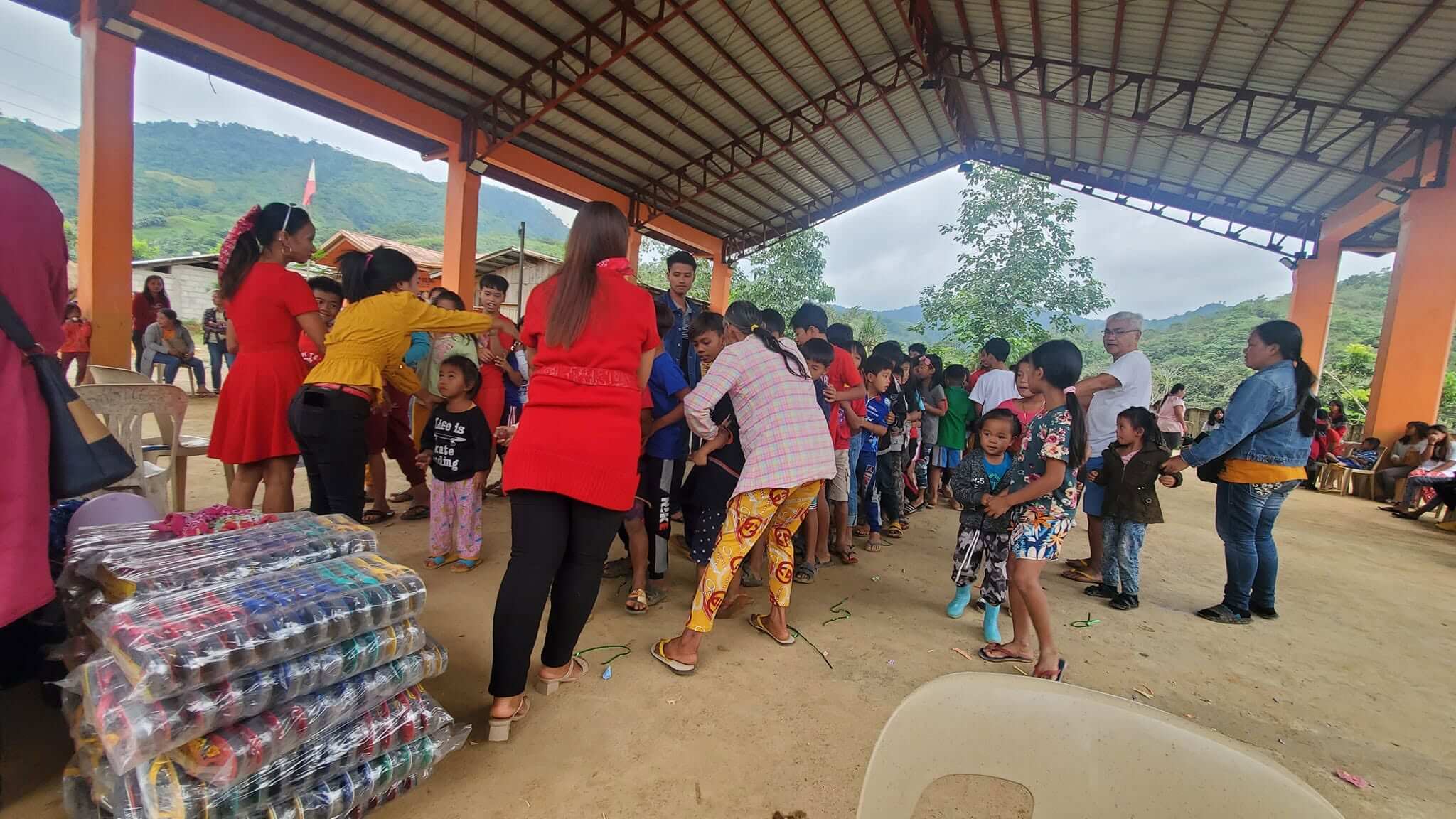 Stacks of packaged slippers piled in the foreground as children and families form a long queue at the distribution area, volunteers managing the line
